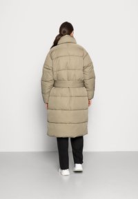 Abrigo acolchado de longitud a la rodilla en color beige con un cuello alto, cintura ceñida y textura acolchada y esponjosa. Combinado con pantalones negros y zapatillas blancas.
