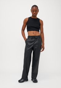 Zwarte crop top gecombineerd met hoge, loszittende zwarte neplederen broek. Model draagt sportieve zwarte schoenen en staat tegen een effen achtergrond.