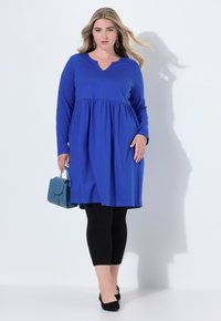 Modré šaty s dlouhým rukávem, V výstřihem a nařaseným pasem, v kombinaci s černými legínami a černými balerínami. Drží malou tyrkysovou kabelku.