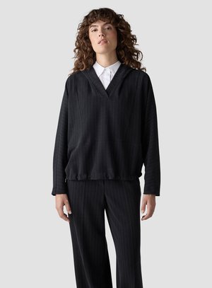 Woman with curly hair wearing a black pinstripe V-neck blouse over a white collared shirt and matching black pinstripe pants.