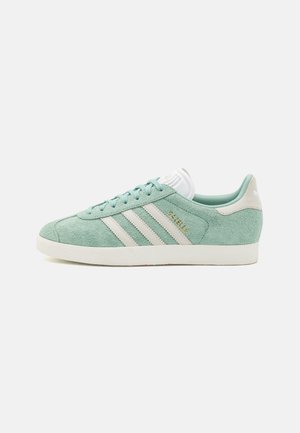 Zapatillas de ante verde claro con rayas y acentos blancos, cuello acolchado y suela de goma texturizada. Logotipo dorado "Gazelle" en el lateral.