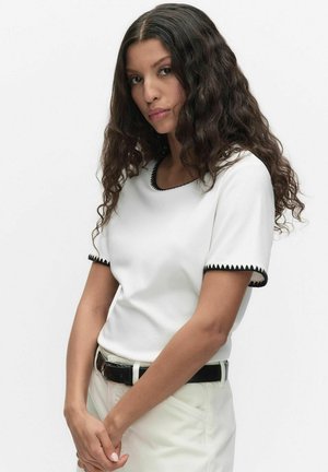 Kvinde med langt krøllet hår iført en hvid kortærmet top med sort kant og lyse bukser med sort bælte, stående mod en hvid baggrund.