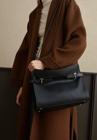 Borsa a tracolla in pelle nera con dettagli in argento, caratterizzata da una texture liscia e doppi manici, appoggiata su un cappotto marrone. Design semplice e rettangolare.