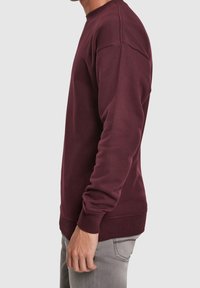 Uomo che indossa una felpa a maniche lunghe bordeaux e jeans grigi chiari, visto di lato su uno sfondo semplice.