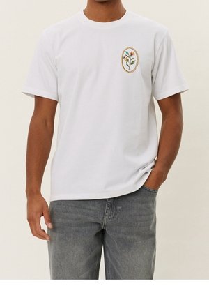 Mann trägt weißes T-Shirt mit kleiner Blumenstickerei auf der linken Brust, kombiniert mit grauer Jeans, Hand in der Tasche.