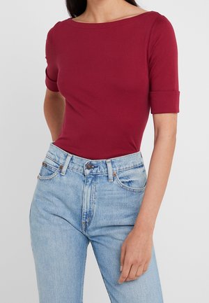 Frau trägt ein eng anliegendes, bordeauxrotes Oberteil mit Ellbogenärmel, das in eine hochtaillierte, hellblaue Jeans mit Fronttaschen und Knopfverschluss gesteckt ist.