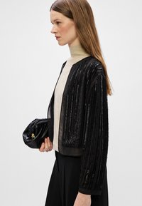 Ελαφρύ μαύρο μπουφάν με κατακόρυφες ρίγες, διάφανο ύφασμα και χαλαρή εφαρμογή. Συνδυασμένο με μπεζ ζιβάγκο και μαύρη τσάντα.