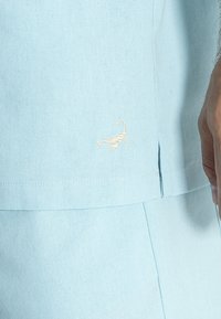 Camicia azzurra a maniche corte realizzata in tessuto testurizzato, con un piccolo scorpione bianco ricamato sul lato inferiore vicino all'orlo.
