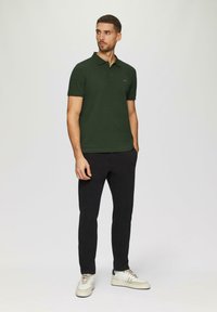 Ciemnozielona koszulka polo o slim fit, z krótkimi rękawami i kołnierzykiem, zestawiona z czarnymi spodniami i białymi trampkami. Prosta szczegółowa grafika na koszulce.