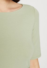 Světle zelený přiléhavý top s krátkým rukávem a kulatým výstřihem, vyrobený z měkké, elastické tkaniny. Má hladkou texturu a minimalistický design.