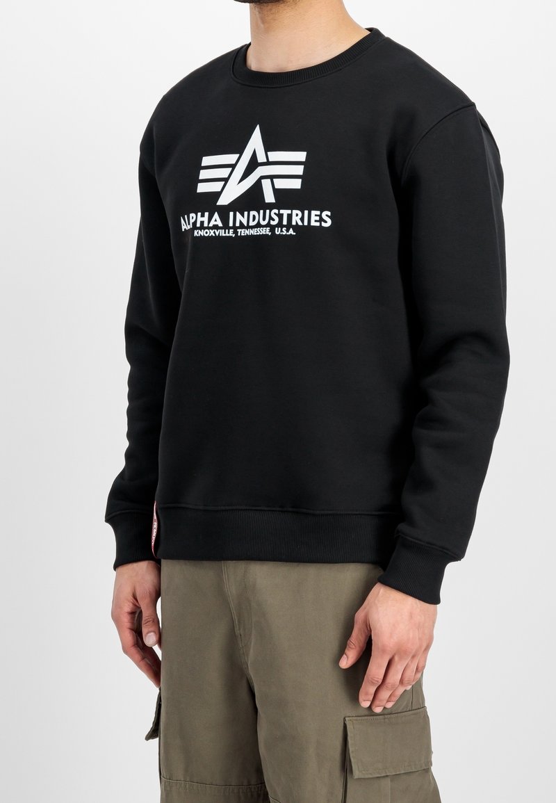 Sweat-shirt noir avec logo Alpha Industries blanc et texte, porté par une personne portant un pantalon cargo olive sur un fond blanc uni.