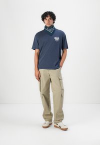 Marinblå t-shirt med grafisk tryck på framsidan, beige cargobyxor med sidofickor, vita sneakers och en mönstrad blå bandana runt halsen.