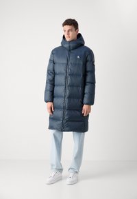 Marineblauer Puffermantel mit Kapuze, ausgestattet mit einem Frontreißverschluss, Seitentaschen und Logo-Detail auf der Brust. Getragen über hellblauen Jeans und weißen Sneakers.