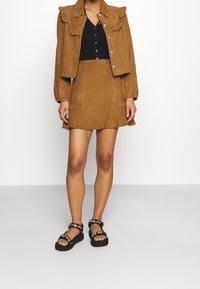 Veste en daim brun avec col à volants, associée à un haut noir boutonné et une mini-jupe en daim brun. Sandales noires avec ferrures décoratives.