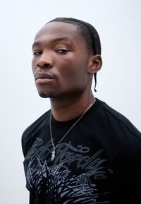 Must musta, millel on hall graafiline disain, kus on keerised ja tekst. Hõbedane kett, millel on ripats. Suurendatud pilt ülakehast ja näost.