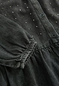 Vêtement gris foncé à manches longues avec de petites perles argentées dispersées sur le tissu, poignet élastique et boutons noirs le long de la couture avant.