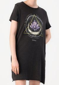 Mujer vistiendo un vestido negro de manga corta con un diseño de loto púrpura, luna creciente, triángulo amarillo y texto motivacional.