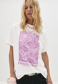 En blond person iført en hvid T-shirt med et lilla kystdesign, der viser sol, bølger, en skildpadde, palmer og teksten "ENDLESS COAST."