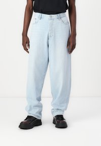 Light blue, loose-fitting jeans made of denim with a straight cut, featuring front pockets and a button closure. Black chunky shoes are worn.
