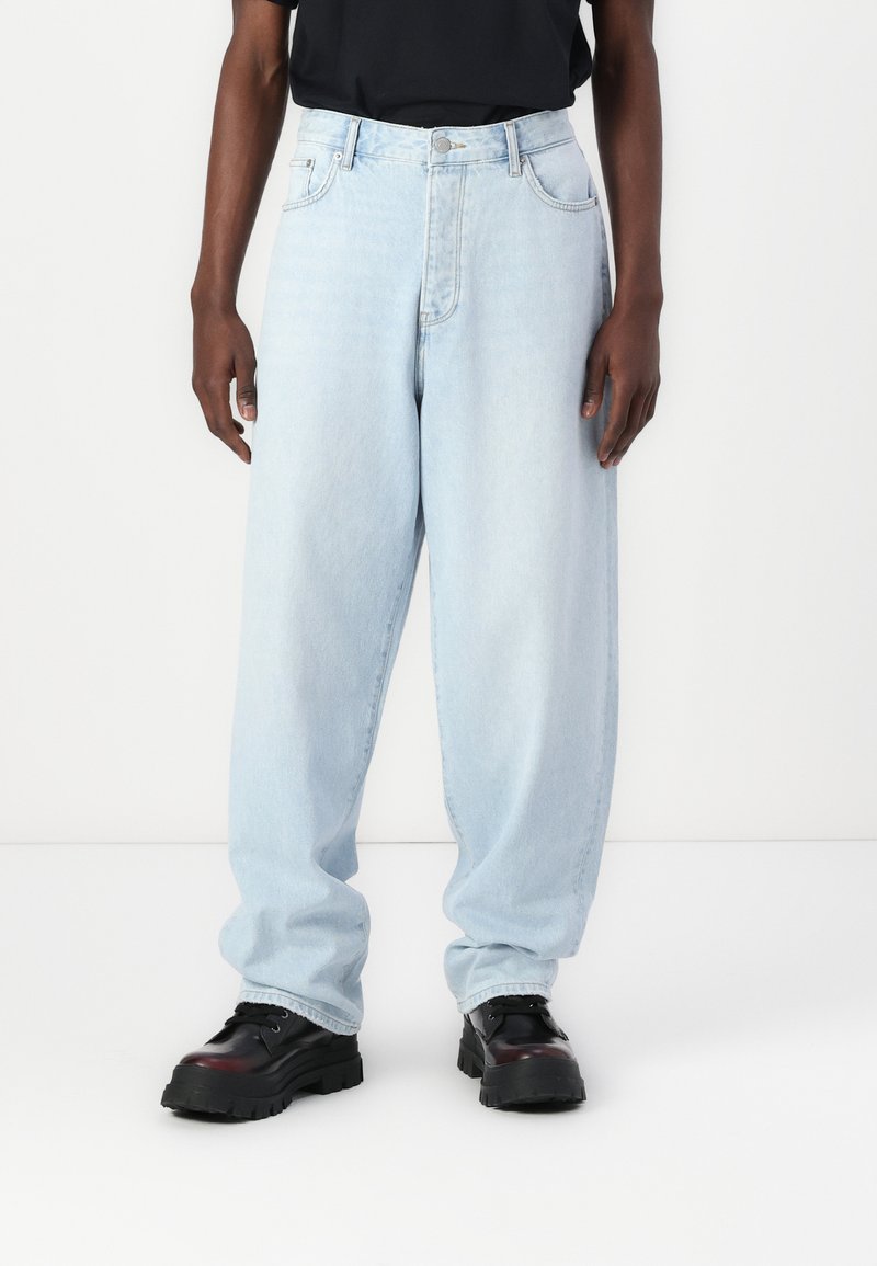 Light blue, loose-fitting jeans made of denim with a straight cut, featuring front pockets and a button closure. Black chunky shoes are worn.