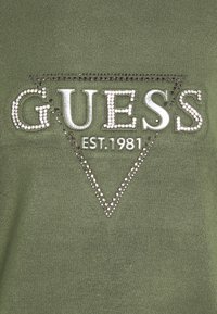 Zielona bluzka z bawełny z domieszką, z napisem "GUESS" w srebrnym kolorze, ozdobionym dżetami, otoczonym trójkątnym wzorem i z napisem „EST. 1981” poniżej.