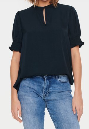 Blouse marine à manches courtes avec un décolleté en forme de trou de serrure et des poignets froncés élastiques, associée à un jean en denim bleu clair.
