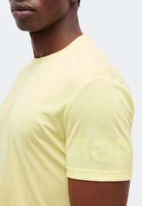 Κοντινό ενός ατόμου που φοράει ανοιχτόκίτρινο κοντομάνικο πουκάμισο με υφασμάτινη υφή και μερικώς ανάγλυφο κείμενο «BOR» στο μανίκι.