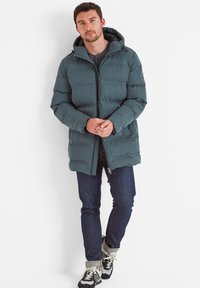Padded dark teal coat with hood, featuring a zip closure and side pockets. Paired with dark jeans and sneakers, standing against a white background.