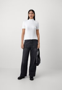 Witte geribbelde top met korte mouwen en een hoge kraag, gecombineerd met loszittende zwarte denim jeans en zwarte schoenen, en een zwarte tas vasthoudend.