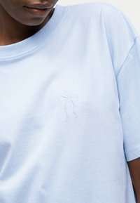 Κοντινό πλάνο σκοτεινόχρωμου ατόμου που φοράει γαλάζιο T-shirt με μικρή κεντημένη λεπτομέρεια φιόγκου στο στήθος, κοιτάζοντας ελαφρώς προς τα κάτω.