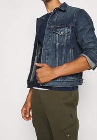 Jeansjacka i mörkblått med en klassisk design, som har två bröstfickor, silverknappar och uppvikta ärmar. Bärs över en marinblå skjorta.