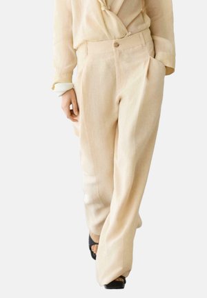 Beige broek met wijde pijpen en plooien, hoge taille, knoopsluiting, gedragen met een licht beige blouse en zwarte open-toe schoenen.