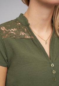 Chemise vert olive avec un panneau en dentelle sur les épaules, manches courtes et un devant à boutons comportant trois boutons. Détail de tissu texturé.