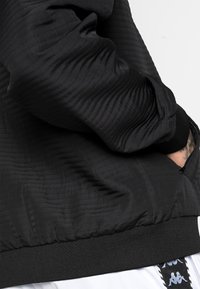 Giacca nera trapuntata con orlo e polsini a costine. Texture liscia con dettagli di tasche con zip. Accentuata da un logo bianco sulla manica.