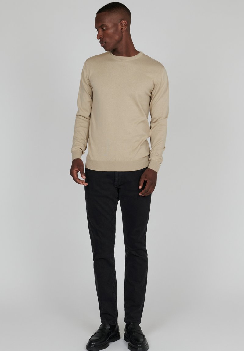 Beige gebreide trui met lange mouwen, ronde hals en geribbelde zoom, gecombineerd met zwarte slim-fit jeans en zwarte schoenen. Eenvoudig en klassiek ontwerp.