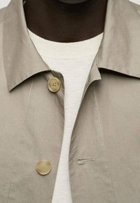 Giacca beige con colletto e due bottoni color cammello, con dettagli di cucitura sottili e una textura morbida, indossata sopra un maglione chiaro.