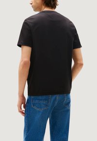 T-shirt nera in cotone con maniche corte, collo rotondo e dettaglio a cucitura a forma di V sul retro, indossata con jeans di denim blu.