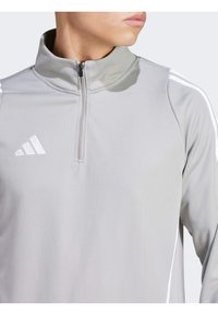 Lichtgrijze kwart-rits sporttop met hoge kraag, voorzien van witte strepen op de schouders en een geborduurd wit logo op de borst.