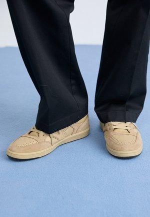 Šviesiai rudos zomšinės sportbačiai su perforuotomis detalėmis ir šviesiai rudos spalvos padais, derinami su juodomis kelnėmis, mėlyname tekstūruotame paviršiuje.