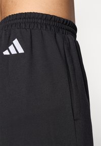 Prim-plan al unor pantaloni atletici negri cu talie elastică, buzunar lateral și un logo triunghiular alb pe partea superioară a coapsei.