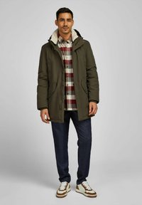 Olijfgroene geïsoleerde parka met een zachte, crèmekleurige bontvoering in de capuchon, gecombineerd met een rood-witte geruite shirt, donkere spijkerbroek en bruine sneakers.