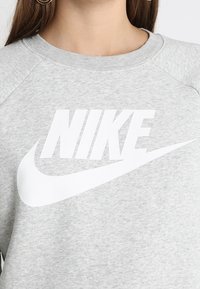 Šedá mikina s velkým bílým logem Nike na hrudi, vyrobená z měkké bavlny, s kulatým výstřihem a raglánovými rukávy.