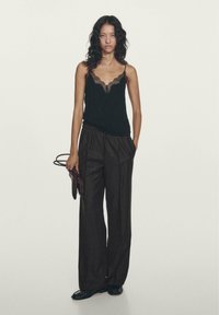 Schwarzes Camisole mit Spitzenbesatz, kombiniert mit weiten Hosen mit dunklen Nadelstreifen. Das Model hält eine braune Handtasche und steht vor einem schlichten Hintergrund.