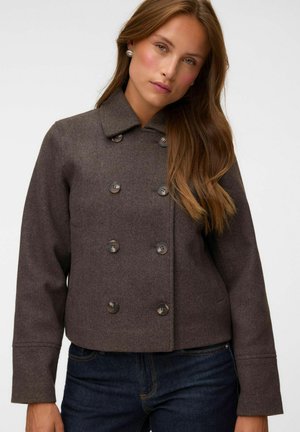 Femme aux longs cheveux bruns portant des boucles d'oreilles en perles, un manteau en laine marron à double boutonnage et un jean bleu foncé, faisant face à un fond uni.