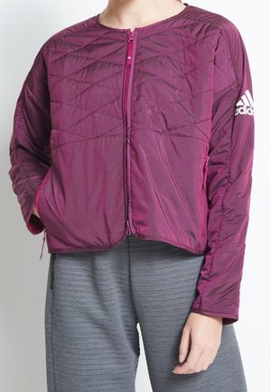 Henkilö pukeutunut tikattuun viininpunaiseen Adidas-takkiin, jossa on etuketju ja logo vasemmassa hihaisessa, yhdistettynä harmaisiin teksturoituihin housuihin.