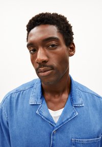 Blauwe spijkerblouse met een kraag, knopen en een borstzak. Soepele textuur en casual ontwerp. Draagt over een wit T-shirt.