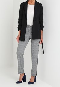 Schwarze Blazer, weißes Oberteil, graue karierte Hose mit Seitenstreifen und blaue Absätze. Der Blazer hat eine taillierte Passform; die Hose verfügt über einen Kordelzug.