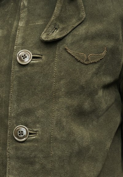 Κοντινή λήψη ενός σακακιού από σουέντ σε ελιά απόχρωση, με δύο κουμπιά, ένα κεντητό έμβλημα σε σχήμα φτερού και μια πλαϊνή τάπα κοντά στον γιακά.