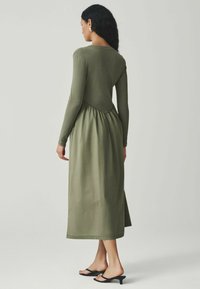 Oliivivihreä mekko, jossa on pitkät hihat, istuva yläosa ja levenevä, kerroksittain rypytetty hame. Sileä pinta ja minimalistinen muotoilu.
