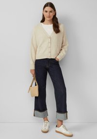 Beige gebreide cardigan met V-hals, knopen aan de voorkant en twee zakken. Gecombineerd met donkere denim jeans en witte sneakers met tan accenten.